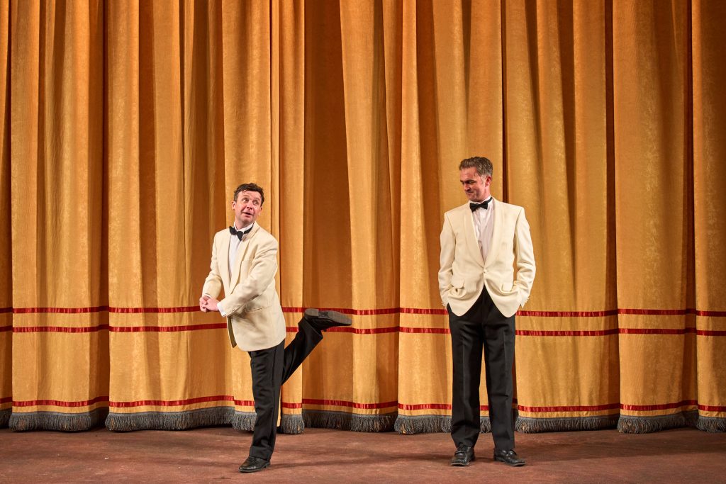 Production photo of two men in front of a theatre curtain