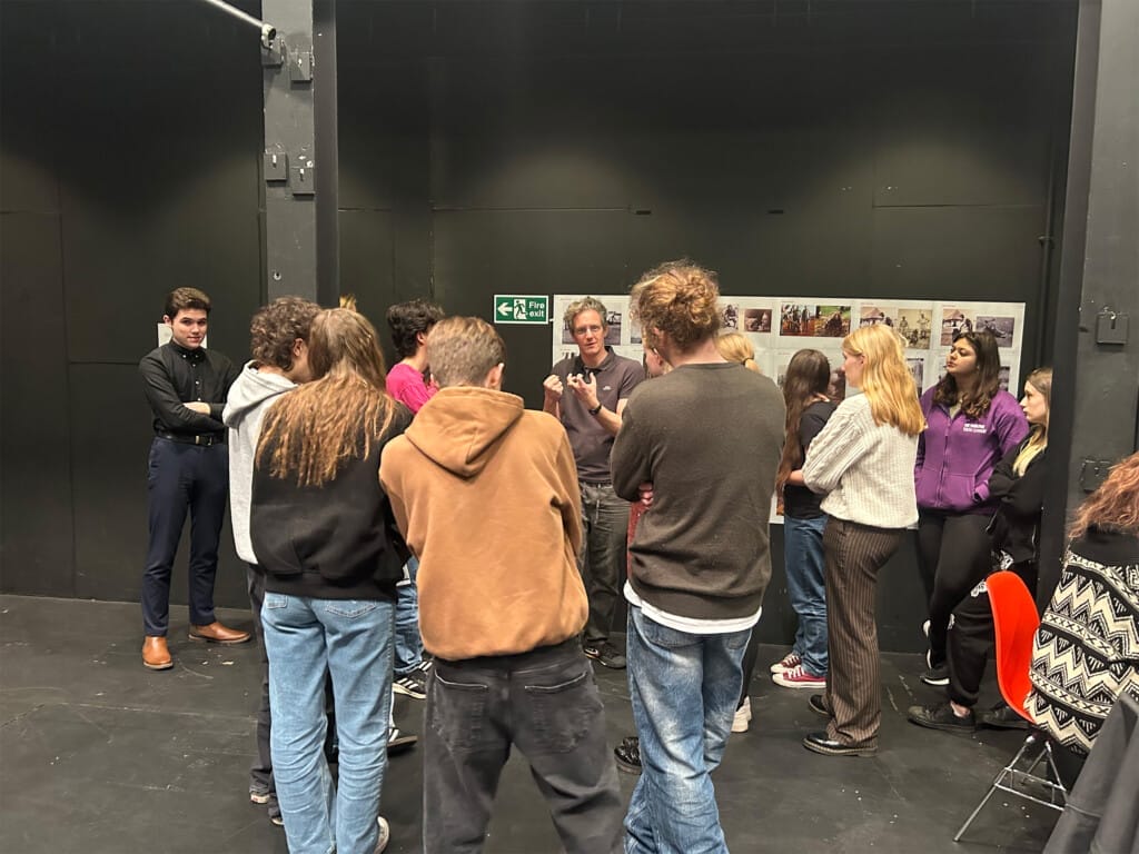 A group of children are standing and listening to the director, who is gestering with his hands.