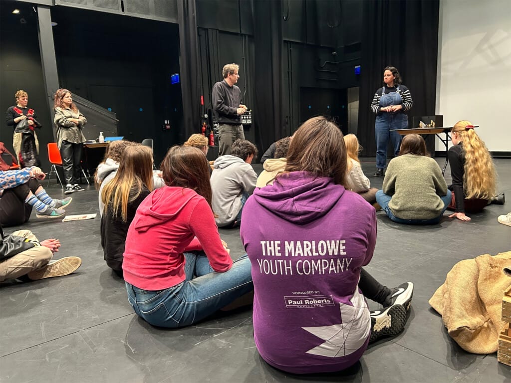 A group of people sit on the floor, listening to a woman standing at the front.