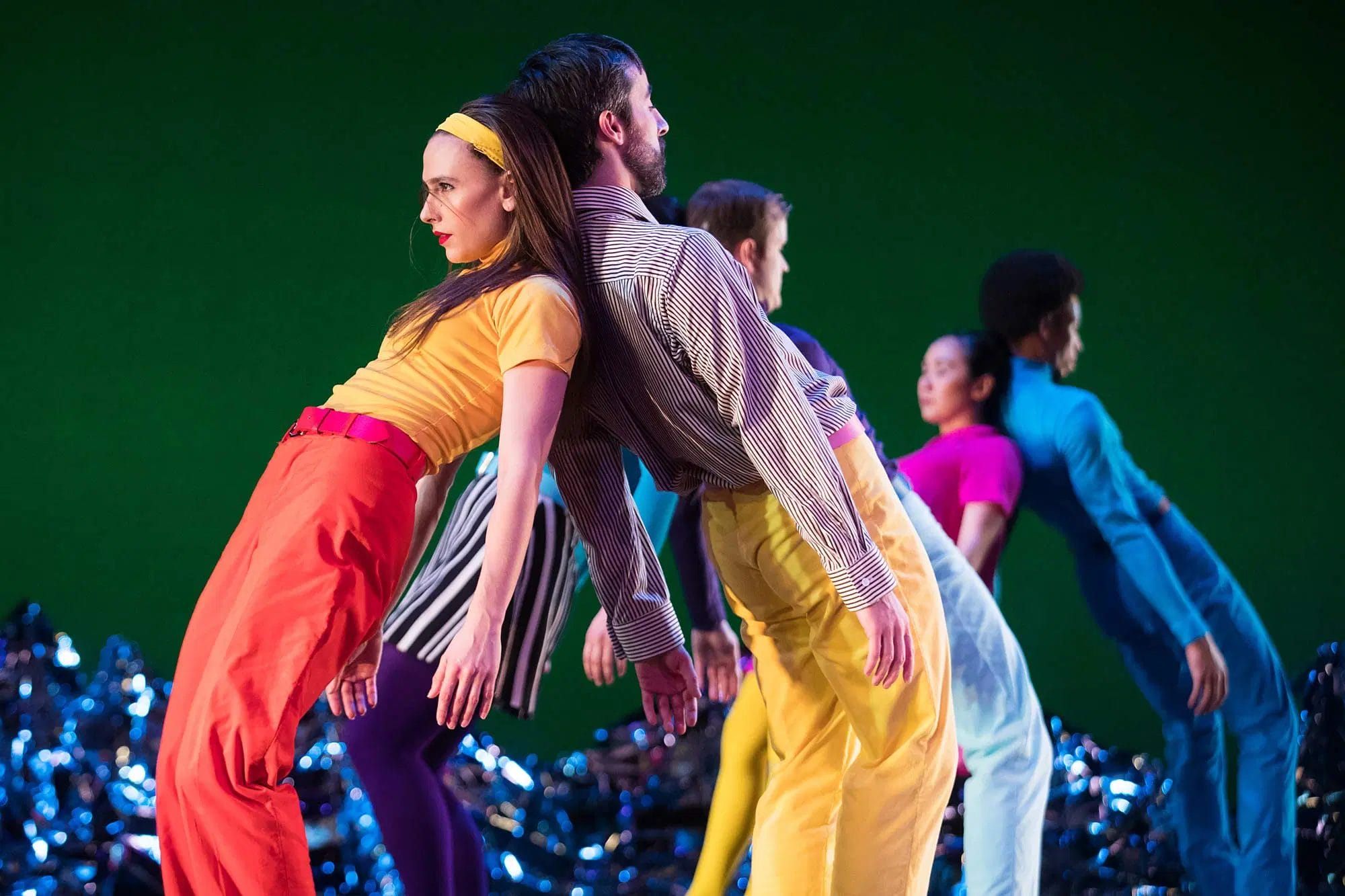 A female dancer wearing red trousers and a yellow top leans back to back with a male dancer wearing yellow trousers and a black and white striped shirt